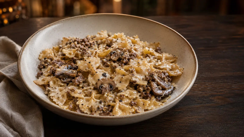 Un tazón de pasta cremosa con carne molida y champiñones, servido sobre una mesa de madera oscura con una servilleta doblada.