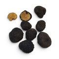 Siete trufas negras, incluidas dos mitades que muestran su interior de color marrón dorado con aspecto de fruto seco, dispuestas sobre un fondo blanco liso.