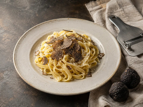 Tagliatelle con virutas de trufa negra, trufas enteras y un rebanador de trufas sobre una encimera de piedra.